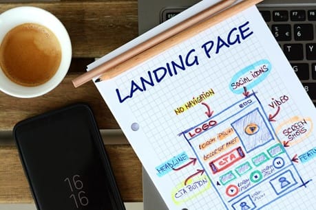 A cup of espresso, a smartphone displaying 16:16, and a notebook with a colorful sketch of a landing page layout lie on a wooden surface next to a laptop keyboard.