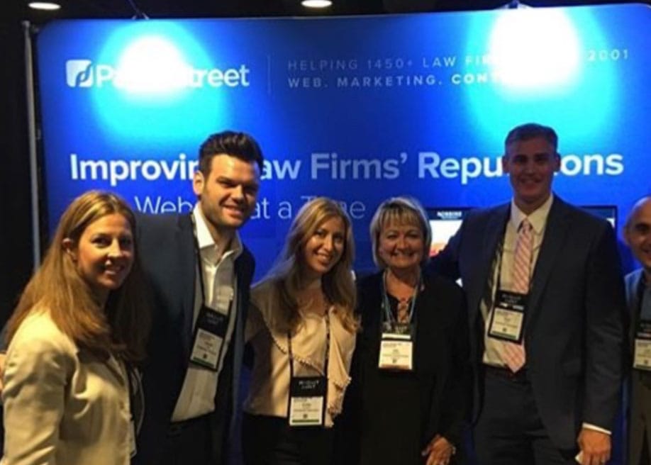 A group of five people standing together in front of a blue display booth that reads "Improving Law Firms’ Reputations" at a professional conference. All are wearing name badges.