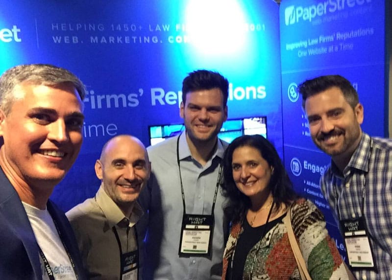 Five people standing together, smiling, posing for a photo at a conference with a blue booth background displaying marketing and law firm-related text.