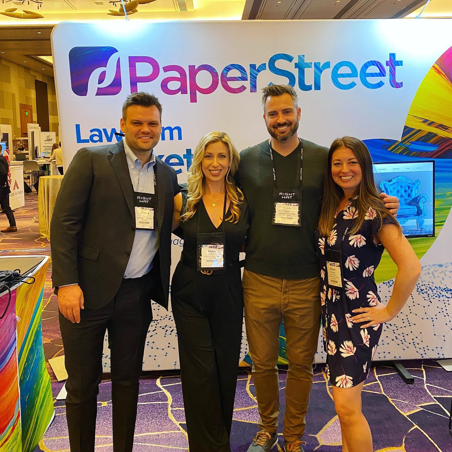 Four people standing together in front of a booth with a "PaperStreet" sign at a convention. They are smiling and wearing name tags.