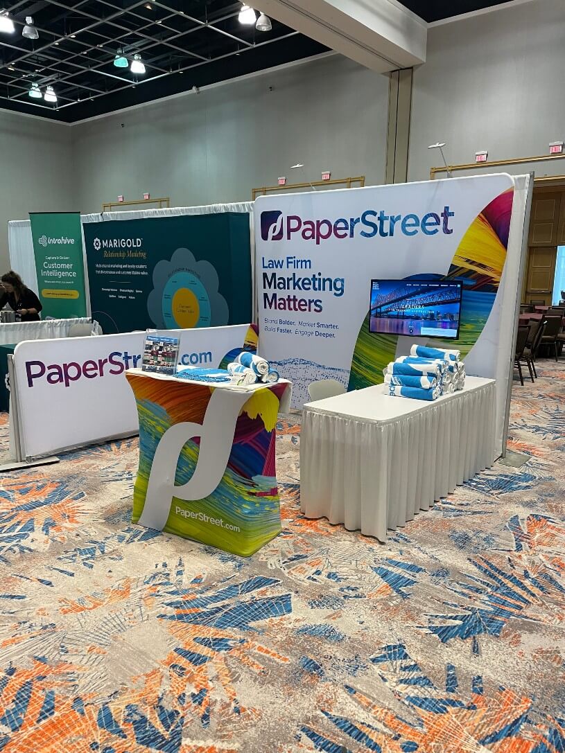 Booths at a convention displaying marketing materials. Prominent booth named "PaperStreet" with a table covered in branded items, a monitor, and a backdrop highlighting "Law Firm Marketing Matters".