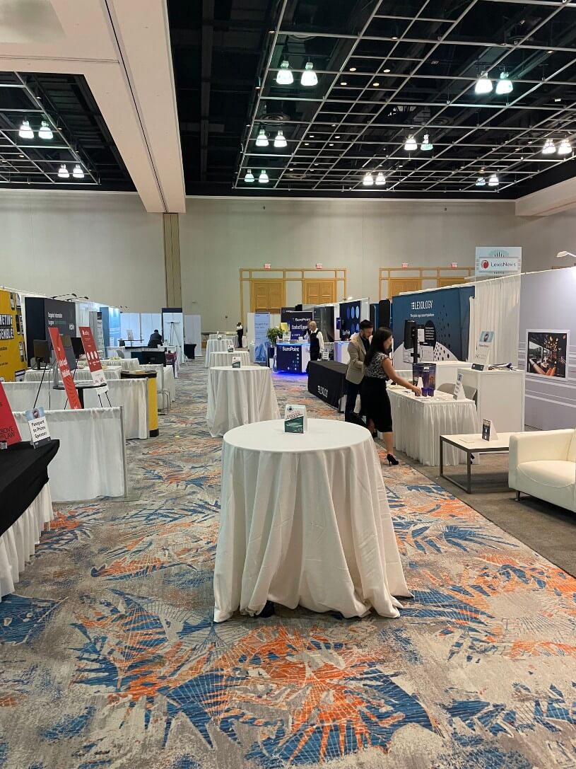 A convention hall with empty tables covered in white tablecloths and various exhibition booths set up along the sides. A few people are standing and interacting at some of the booths.