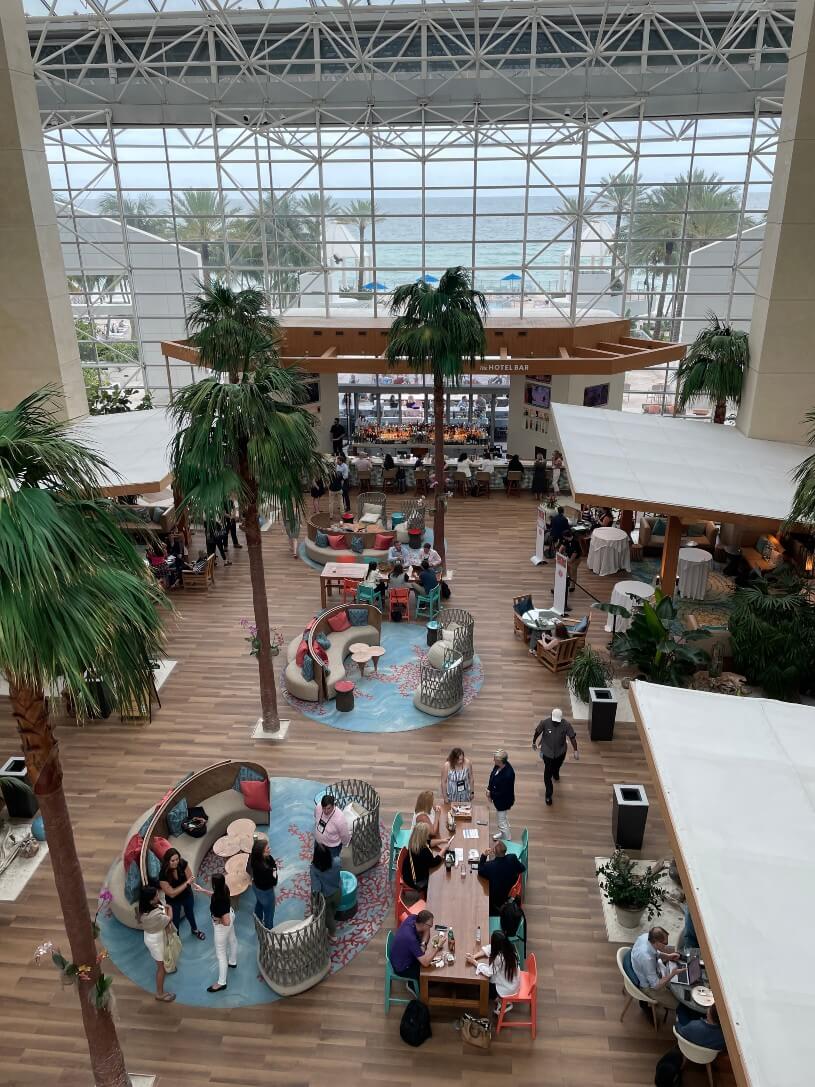 A spacious indoor atrium with large windows, palm trees, and people sitting at tables. The ocean and palm trees outside are visible through the windows.