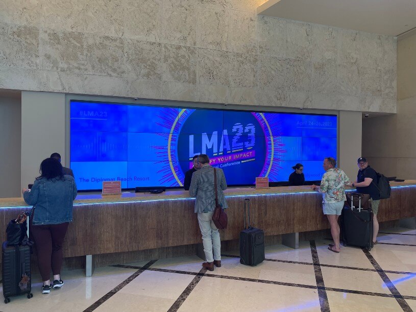 People checking in at a hotel reception desk with large electronic screens displaying "LMA'23 - Elevate Your Impact". Some have suitcases, and staff are assisting them.