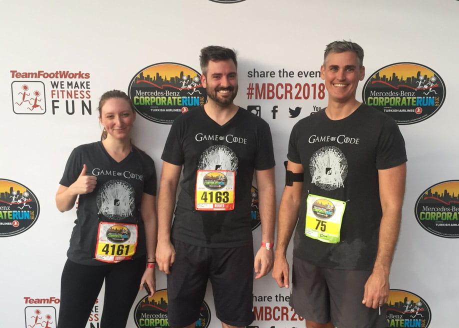 Three people wearing "Game of Code" shirts stand together after a run, smiling and sweating. They have race bibs numbered 4161, 4163, and 75. A branded event backdrop is behind them.