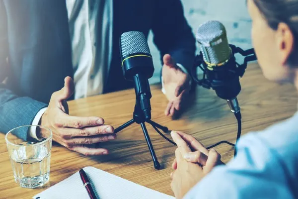 Two people sit at a table with two microphones between them, engaged in conversation. A glass of water, a notepad, and a pen are also on the table.