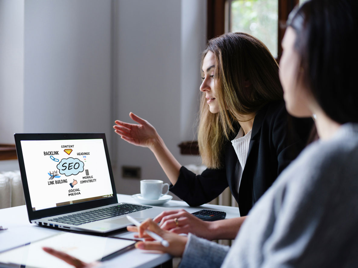 Two women are looking at a laptop screen displaying a diagram about SEO, which includes terms like content, backlinks, social media, and keywords. One woman gestures toward the laptop.