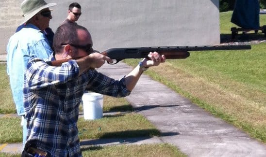 A person stands outdoors aiming a shotgun at a shooting range, with two other individuals standing in the background wearing sunglasses and hats.