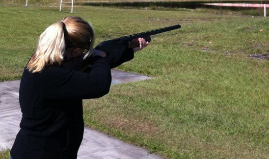 Person with blonde hair in a ponytail, wearing a black jacket, aiming a shotgun outdoors on a grass field.