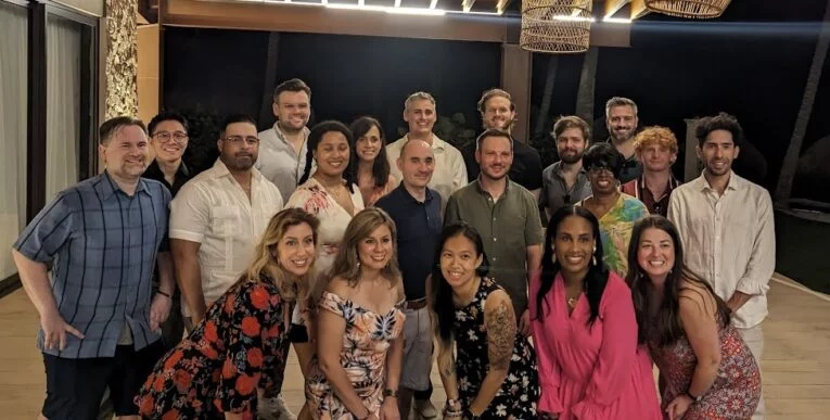 A group of people poses for a photo indoors, many smiling and wearing casual attire. The setting appears to be a well-lit room with a night-time background.