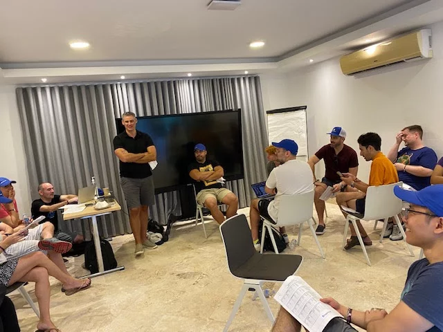 A group of people are seated and standing in a conference room, engaging in a discussion. Some are wearing blue caps and looking at papers, while one person stands and speaks. A TV and flip chart are in the background.