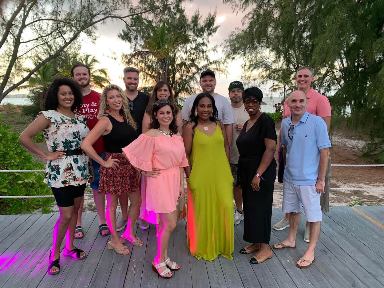 A group of twelve people standing on a wooden deck outdoors with trees and a beach visible in the background. Some are smiling, and they are dressed in casual and semi-formal attire.