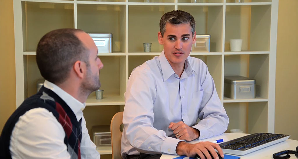 Two men are seated at a table, with one using a laptop while the other listens. They appear to be in conversation, with shelves containing various items in the background.