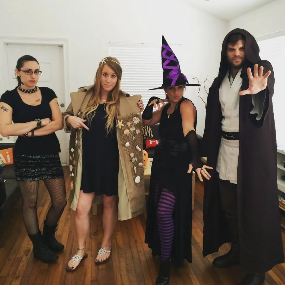 Four adults in a room wearing costumes: a person with glasses and black dress, a person in a cloak with leaves, a person in a witch outfit, and a person in a hooded cloak with an outstretched hand.