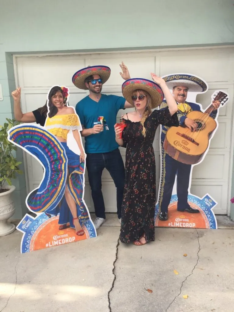 Three people wearing sombreros pose with cardboard cutouts of musicians outside a building, holding drinks and making celebratory gestures.