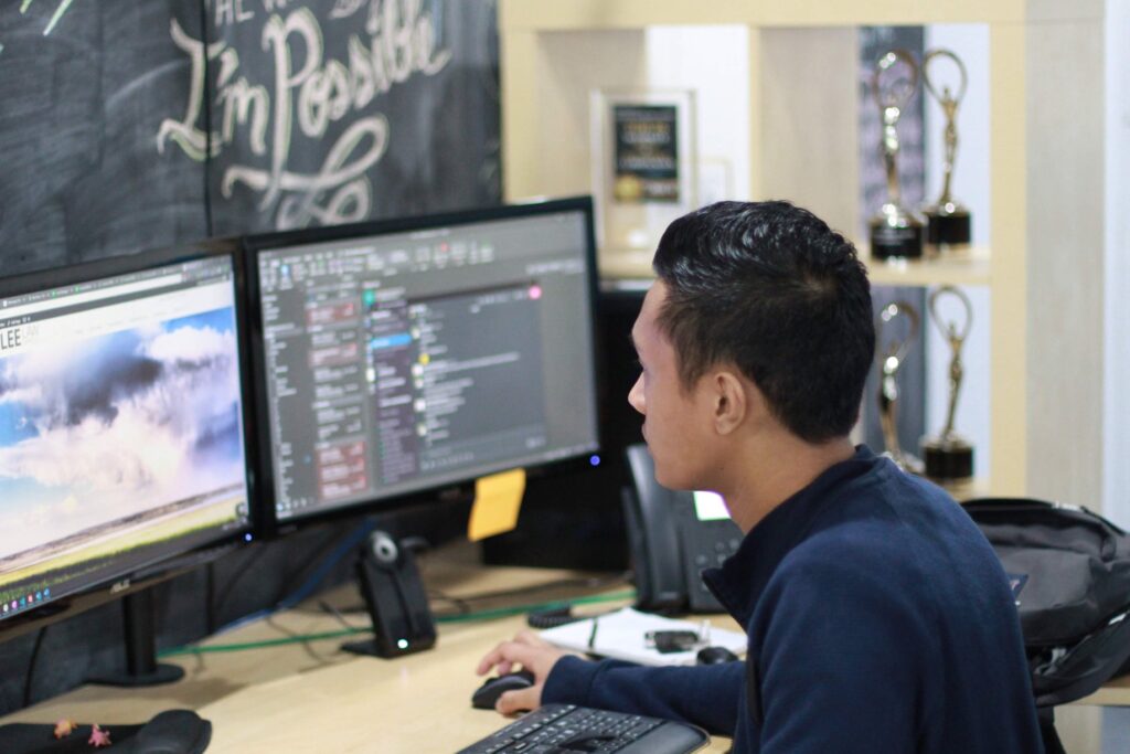 A person works at a desk with two computer monitors, one displaying code and the other an image of clouds. Trophies are visible on a shelf in the background.