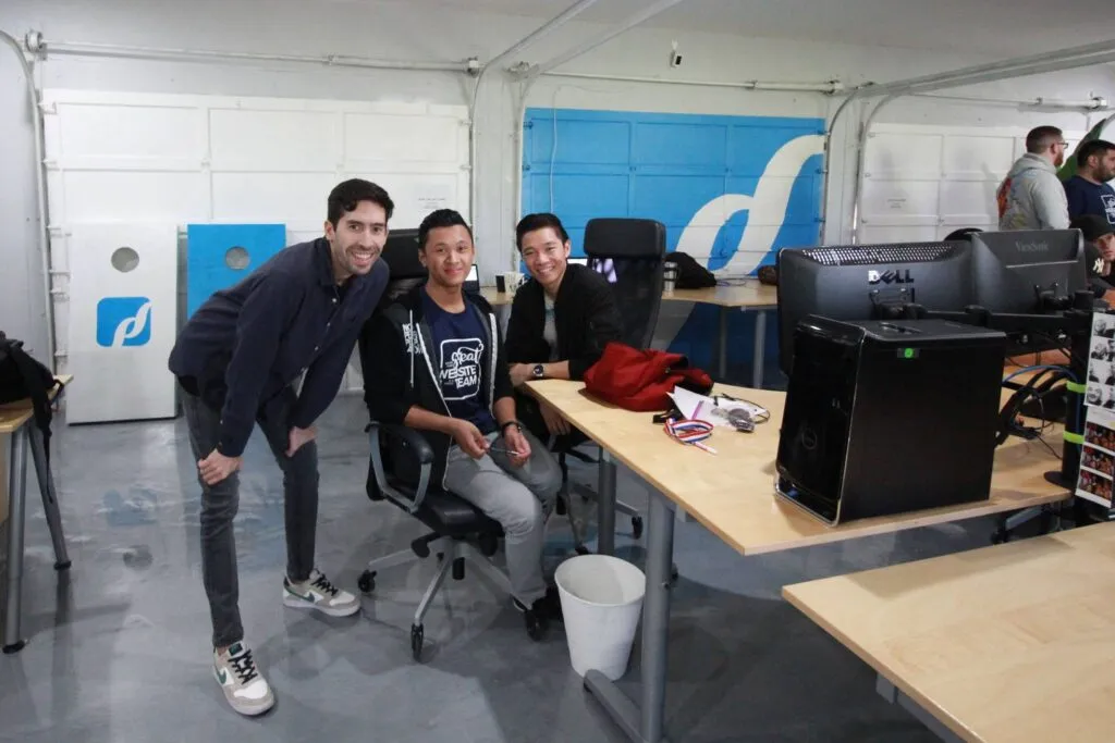 Three people smile at the camera in a tech office space, with computers and workstations visible. One person is seated while two others stand beside him.