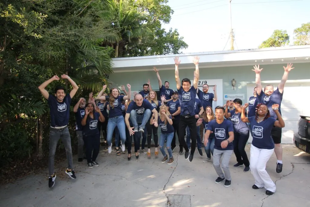 A group of people wearing matching shirts jumps in the air outside a building with a sign that reads "219." Trees and parked cars are visible in the background.