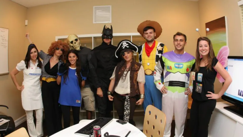 A group of ten people dressed in various Halloween costumes stand together in an office setting.