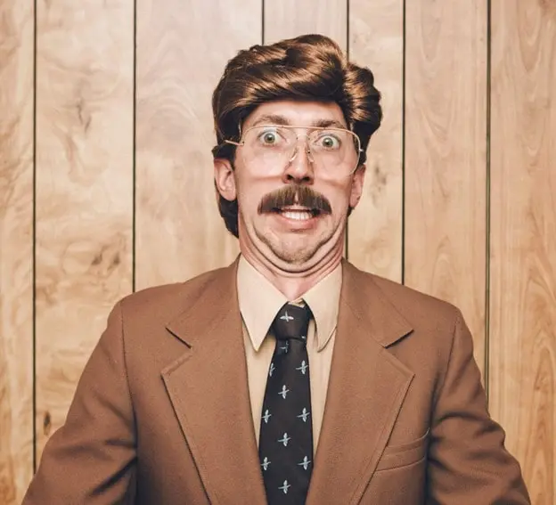 A man wearing a brown suit, tie, and glasses makes a wide-eyed, humorous expression standing against a wood-paneled wall.