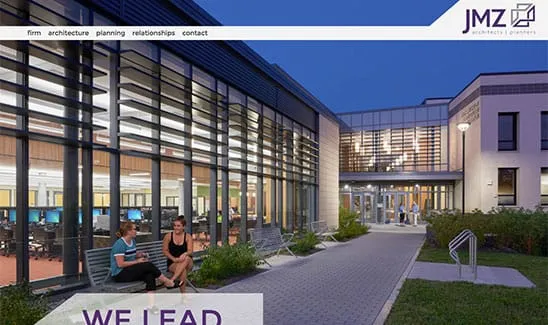 Two people sit on benches outside a modern building with large windows and a walkway leading to the entrance. The building is illuminated from within, and the text “WE LEAD” is partially visible.