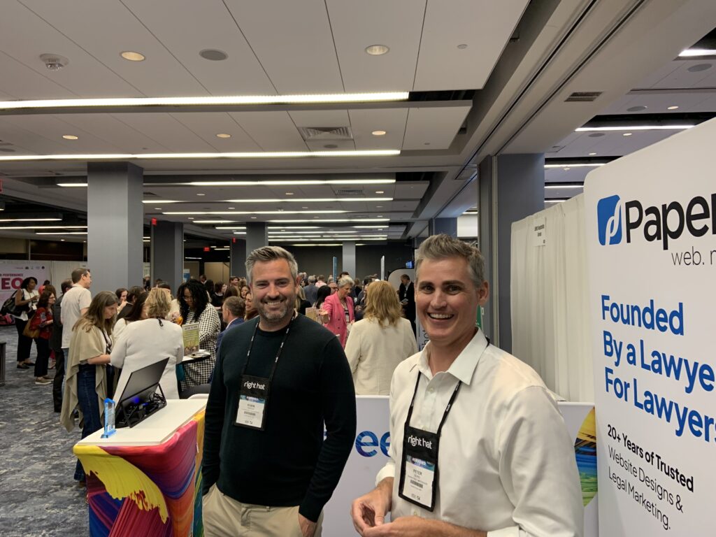 Two men stand at a booth during a busy conference or networking event indoors.