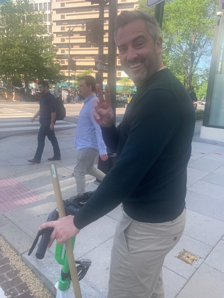 Man smiling and flashing a peace sign while holding the handlebars of a scooter on a city sidewalk.