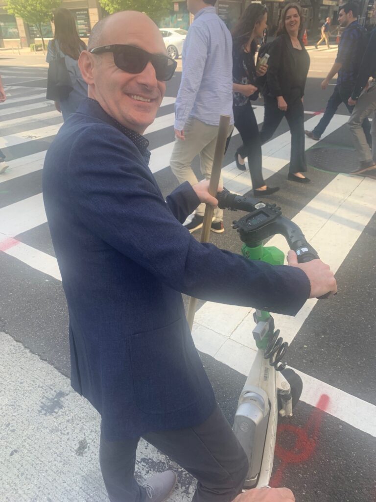 Man in sunglasses and blazer stands with electric scooter at city crosswalk.