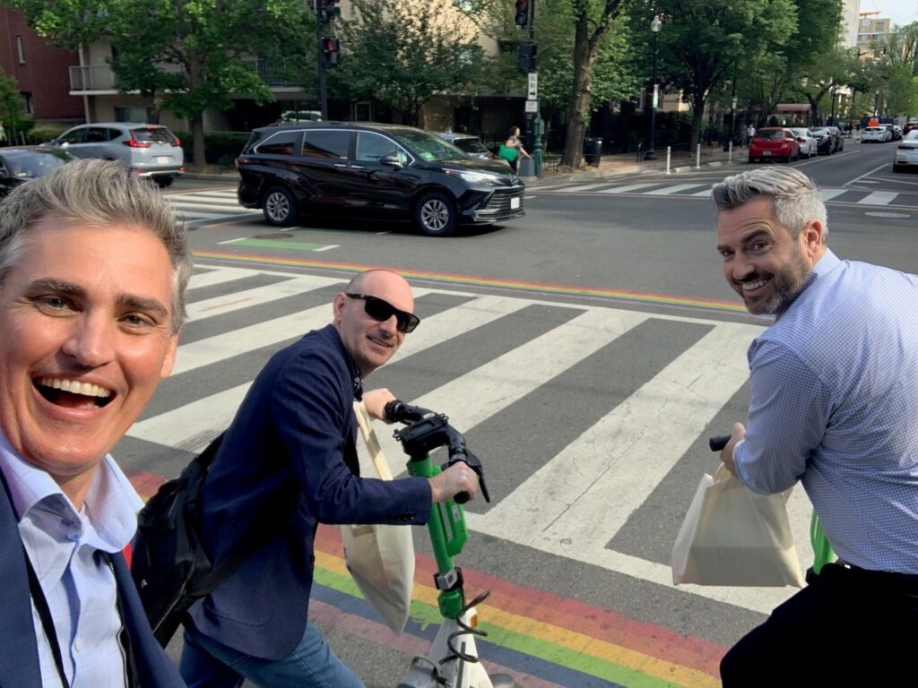 Three people smile while crossing a street with rainbow stripes, two riding electric scooters.